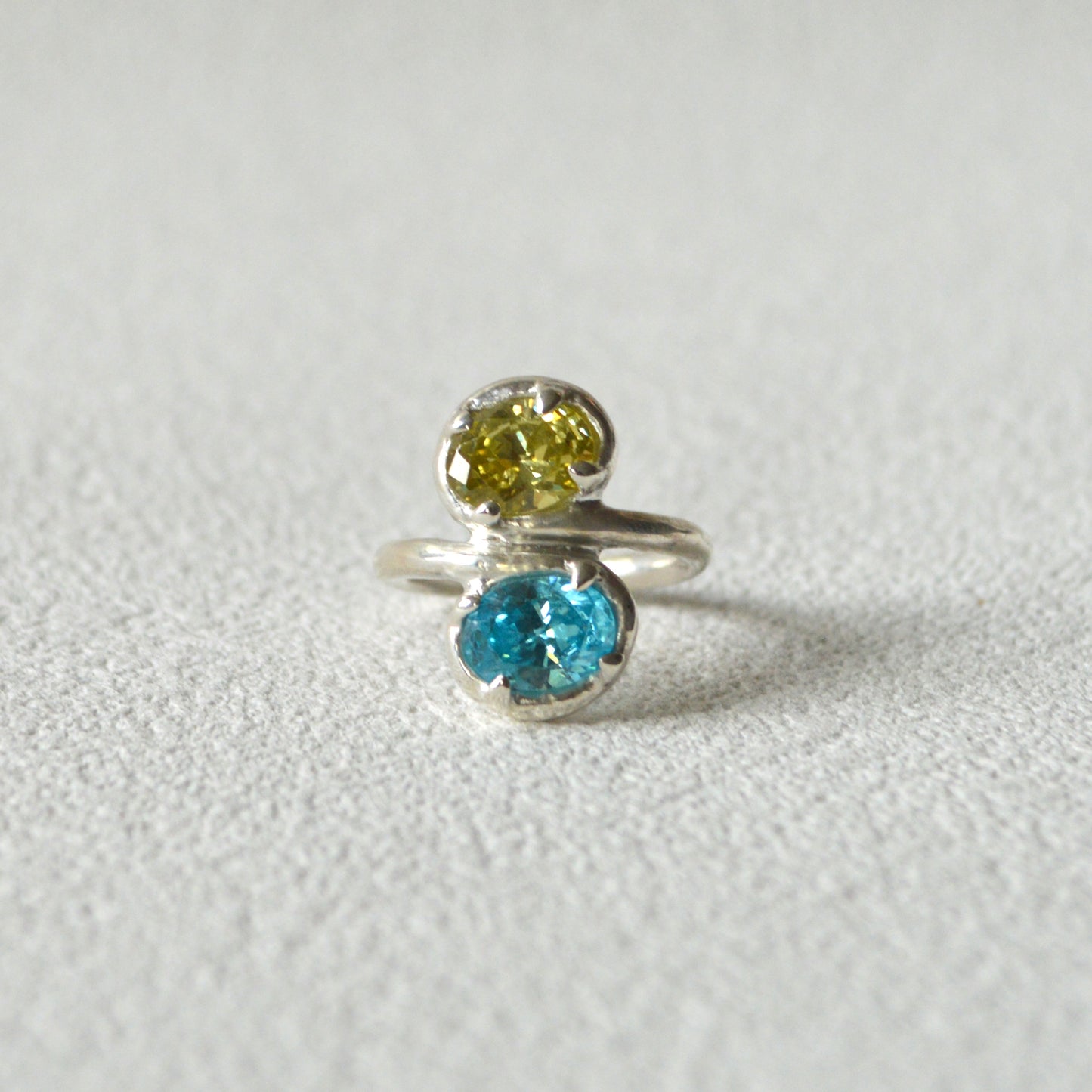 Silver ring with two gemstones, one yellow and one blue, on a light gray background