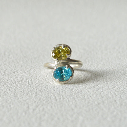 Silver ring with two gemstones, one yellow and one blue, on a light gray background