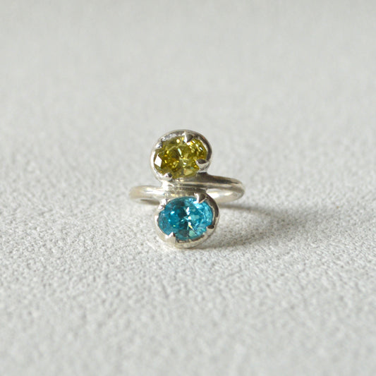 Silver ring with two gemstones, one yellow and one blue, on a light gray background
