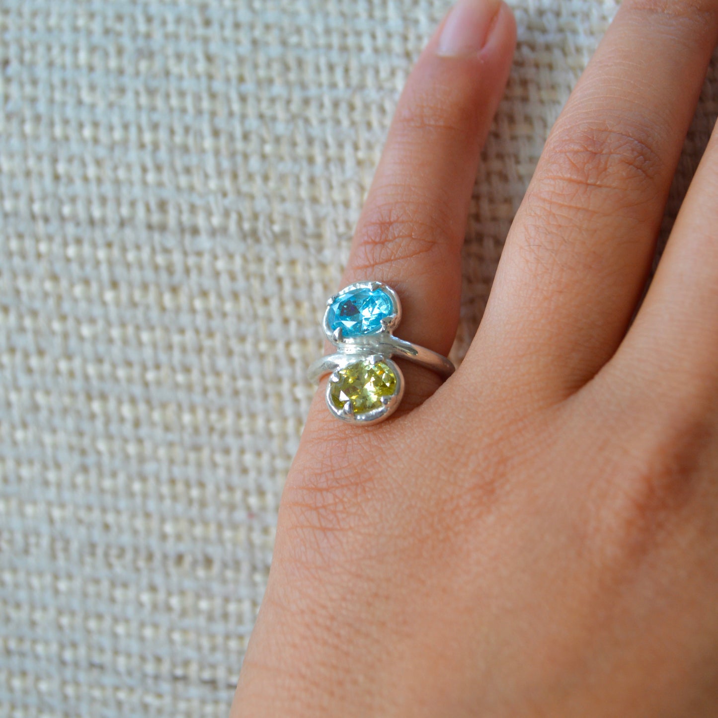 Silver ring with blue and green gemstones on a hand against a textured fabric background
