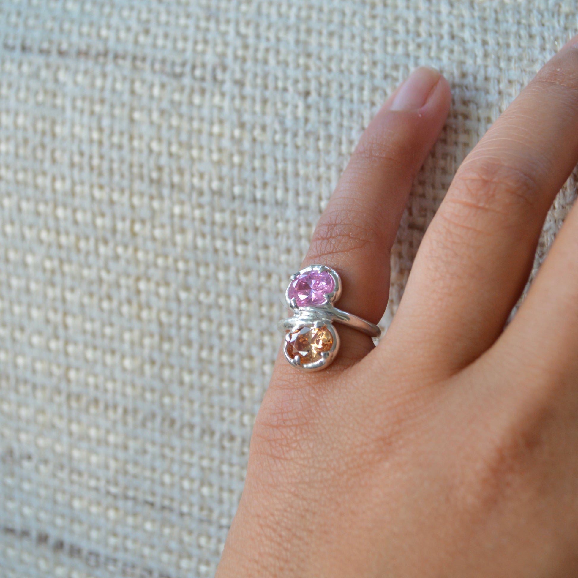 Silver ring with pink and orange gemstones on a hand against a textured beige background