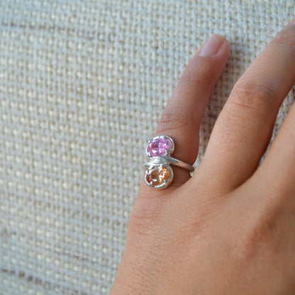 Silver ring with pink and orange gemstones on a hand against a textured beige background